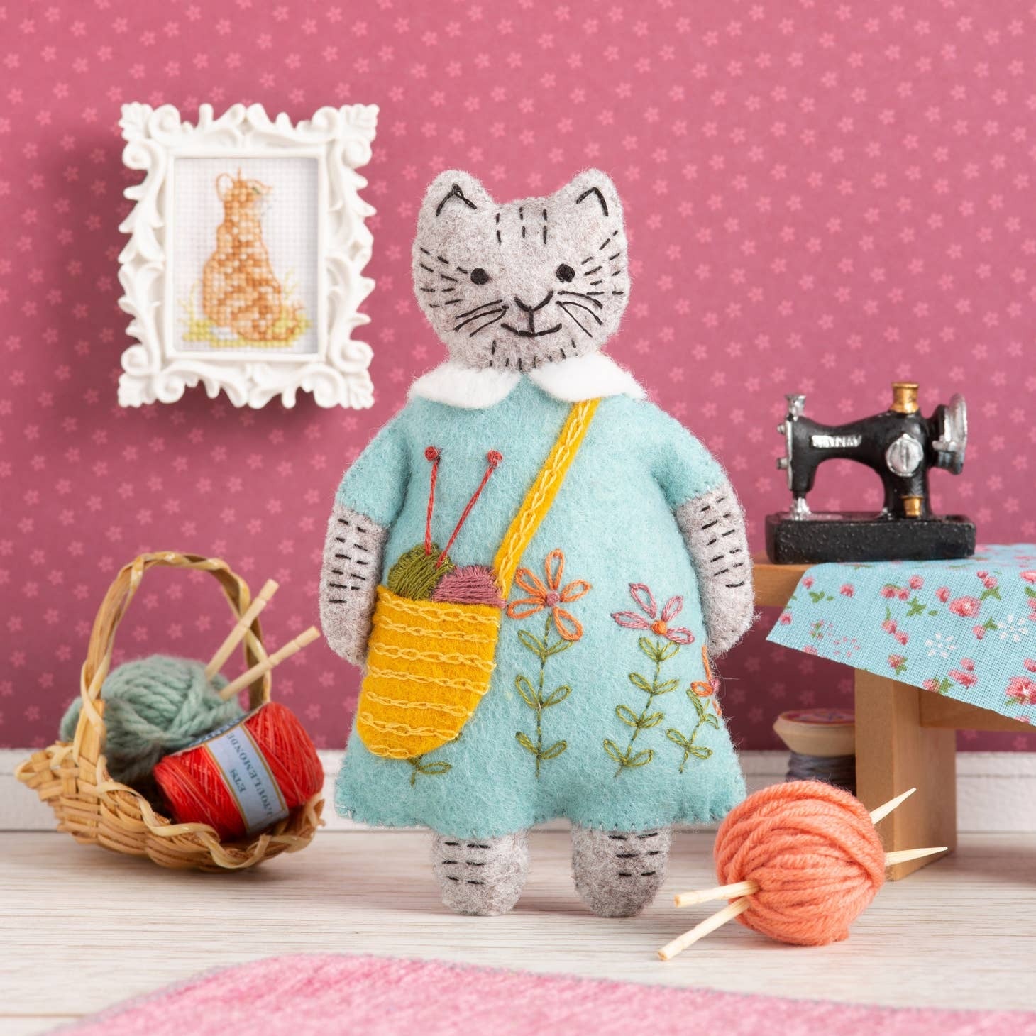 Handcrafted felt cat figure from a DIY felt cat craft kit in aqua dress, knitting with vibrant yarn, set against a pink backdrop with miniature sewing accessories and framed cross-stitch artwork.