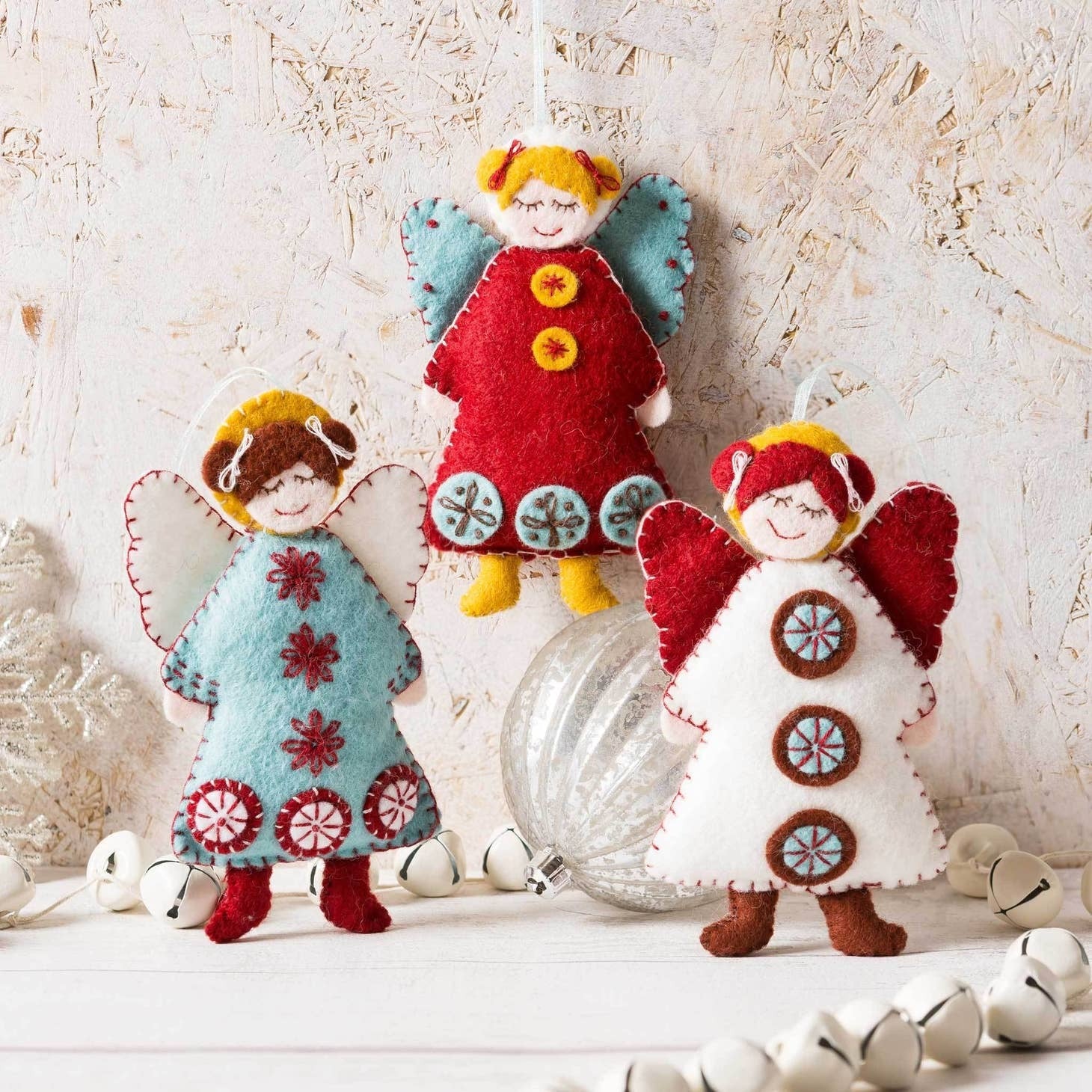 Handcrafted Scandinavian felt angels in vibrant red and blue hues, adorned with intricate embroidery and festive motifs, set against a rustic white background with silver holiday ornaments.