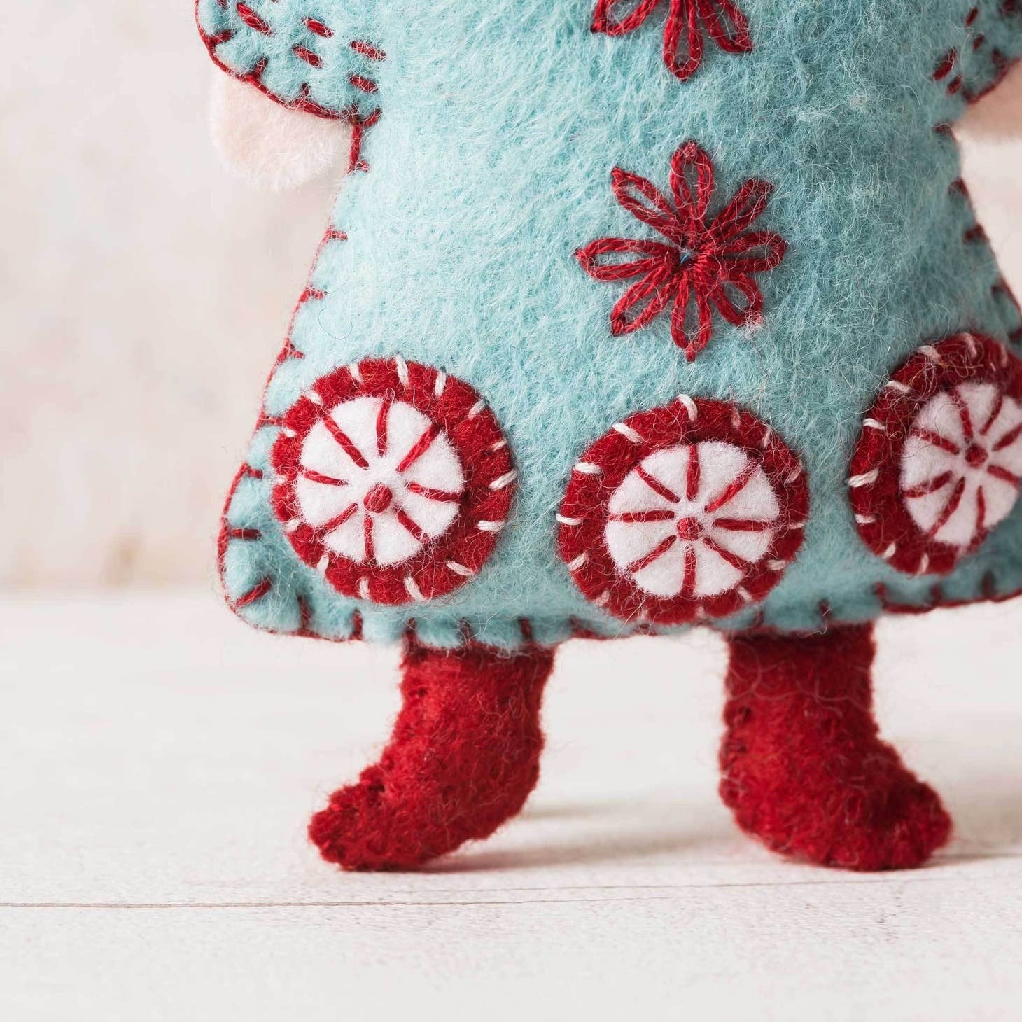 Detail shot of a handcrafted felt angel ornament highlighting the aqua-blue dress adorned with red embroidered snowflakes and plush peppermint candy designs, paired with cozy red felted boots.