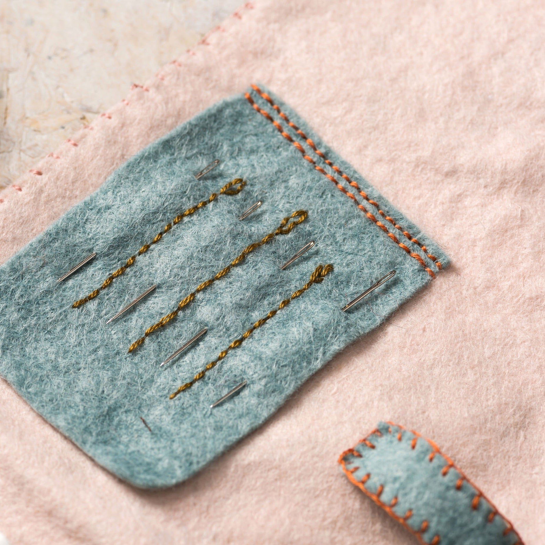 Close-up of a teal felt sewing pocket with intricately stitched orange borders, holding an array of gold-threaded needles, set against a soft pink felt backdrop.