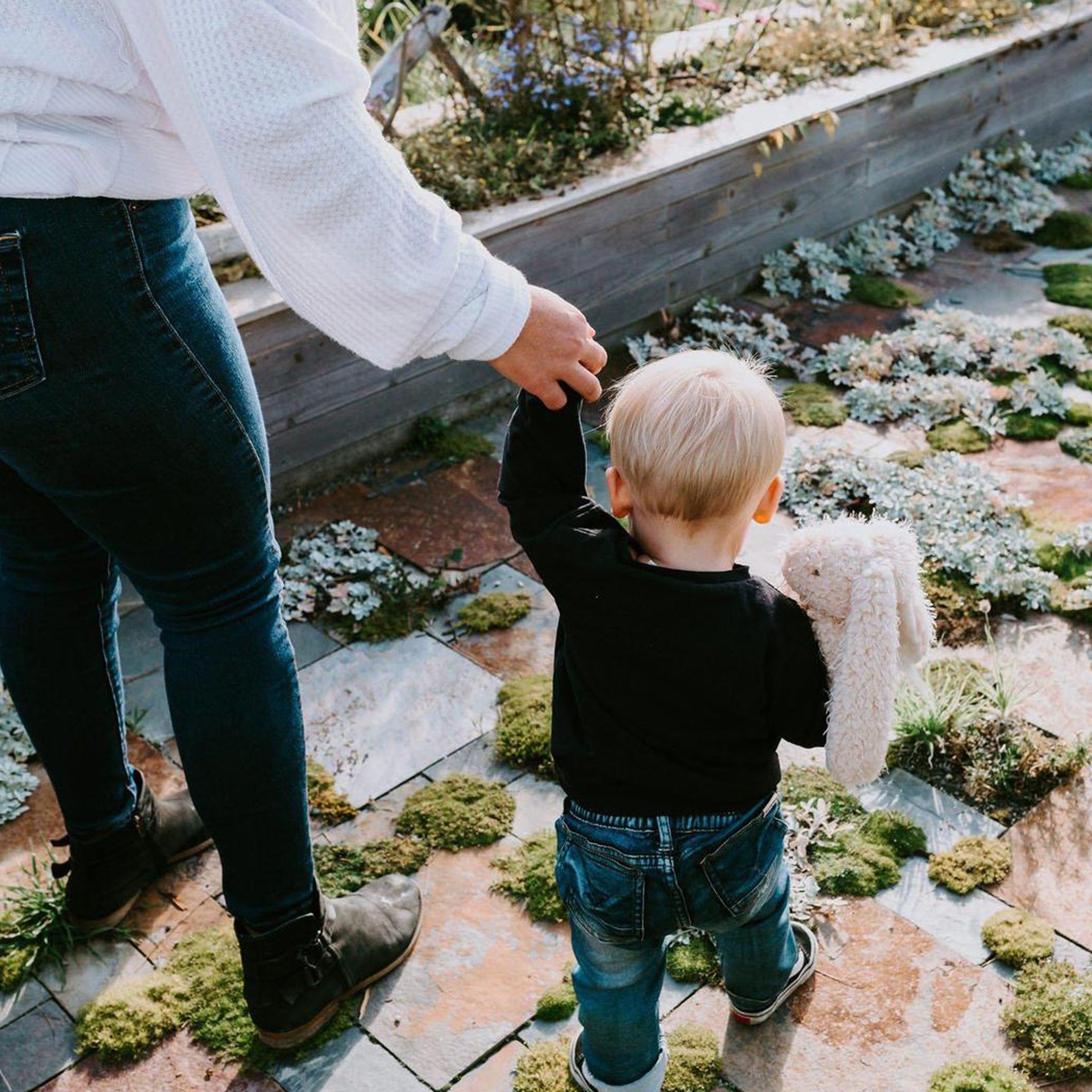 A toddler walking hand-in-hand with an adult, holding Harey the Plush Rabbit, highlighting the rabbit's bushy white tail and vintage charm.
