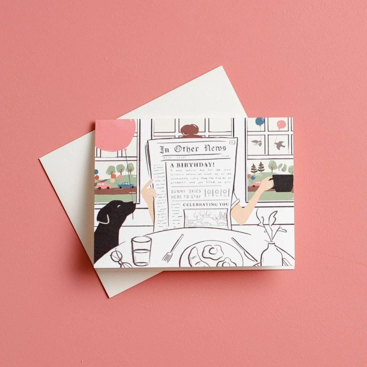 Illustrated chic birthday card on warm white premium cardstock, featuring a woman reading a newspaper at a brunch table with a black lab holding a pink balloon, and a scenic backdrop of birds with balloons through a window.