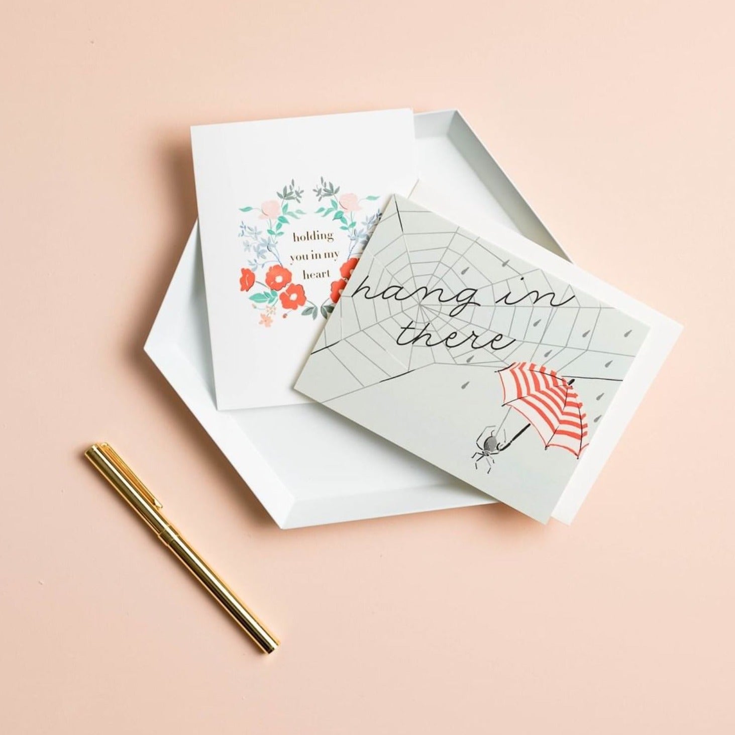 Illustrated sympathy card on premium warm white cardstock, sitting on a white ceramic tray on a peach background, featuring a spider web with the words 'Hang in There' and a spider holding a red and white striped umbrella on a pale blue background.