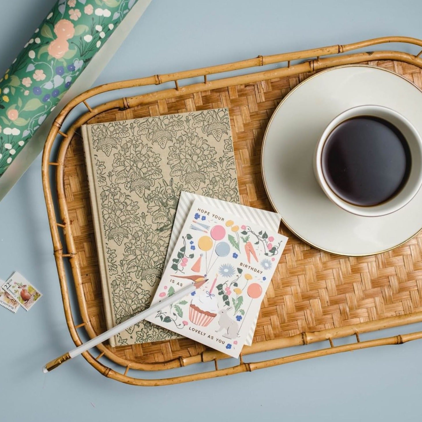 Illustrated 'As Lovely As You' Birthday Card displayed on a rattan tray alongside a floral notebook, white pencil, and coffee cup, featuring a vibrant mix of cupcakes, balloons, and garden florals on warm white cover cardstock.