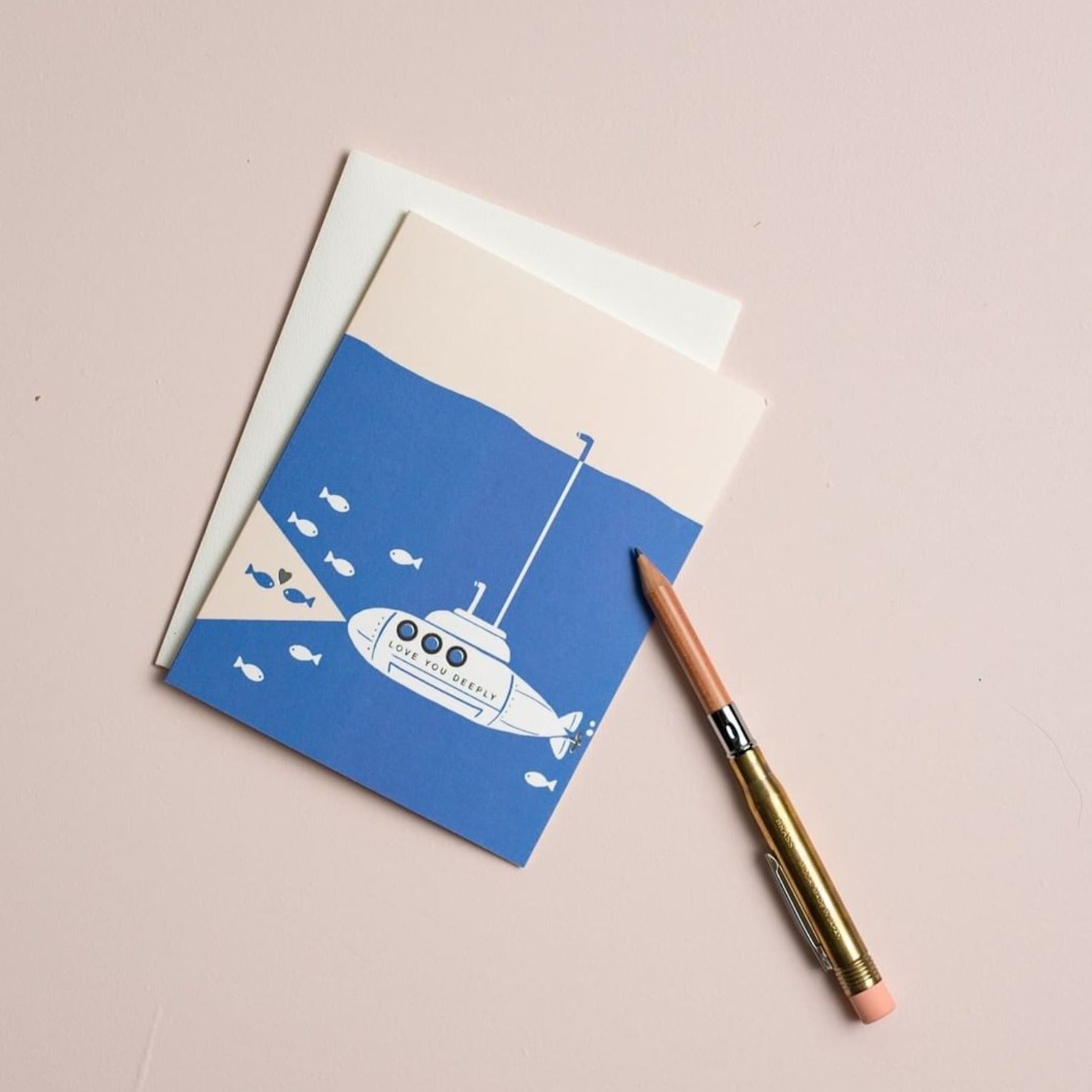 Illustrated love card sitting on a beige background next to a gold pen, with the card printed on warm white premium cardstock, featuring a submarine underwater with fish, and two fish in the submarine's headlight beams sharing a heart, with the text 'Love You Deeply.