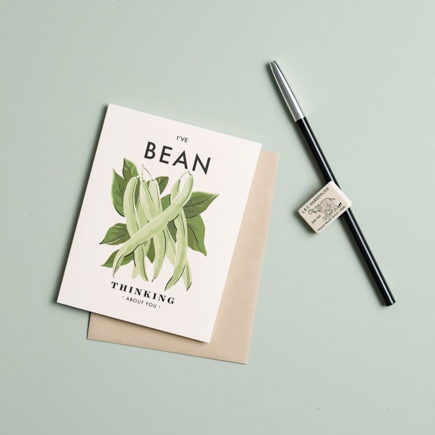 Illustrated 'Thinking About You' card sitting on a pale green background next to a pen and eraser, the card featuring a bunch of beans and the text 'I've Bean Thinking About You.'