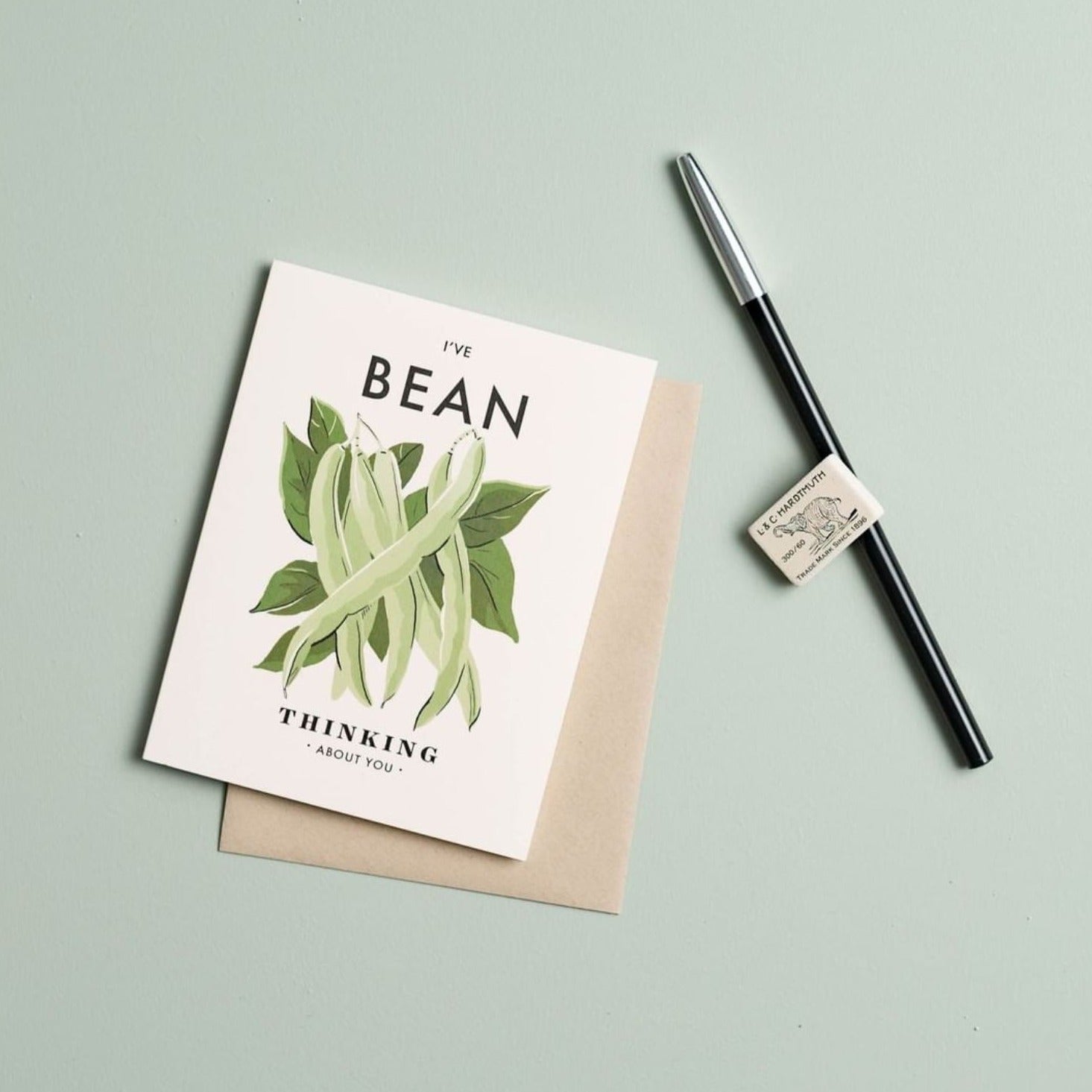 Illustrated 'Thinking About You' card sitting on a pale green background next to a pen and eraser, the card featuring a bunch of beans and the text 'I've Bean Thinking About You.'