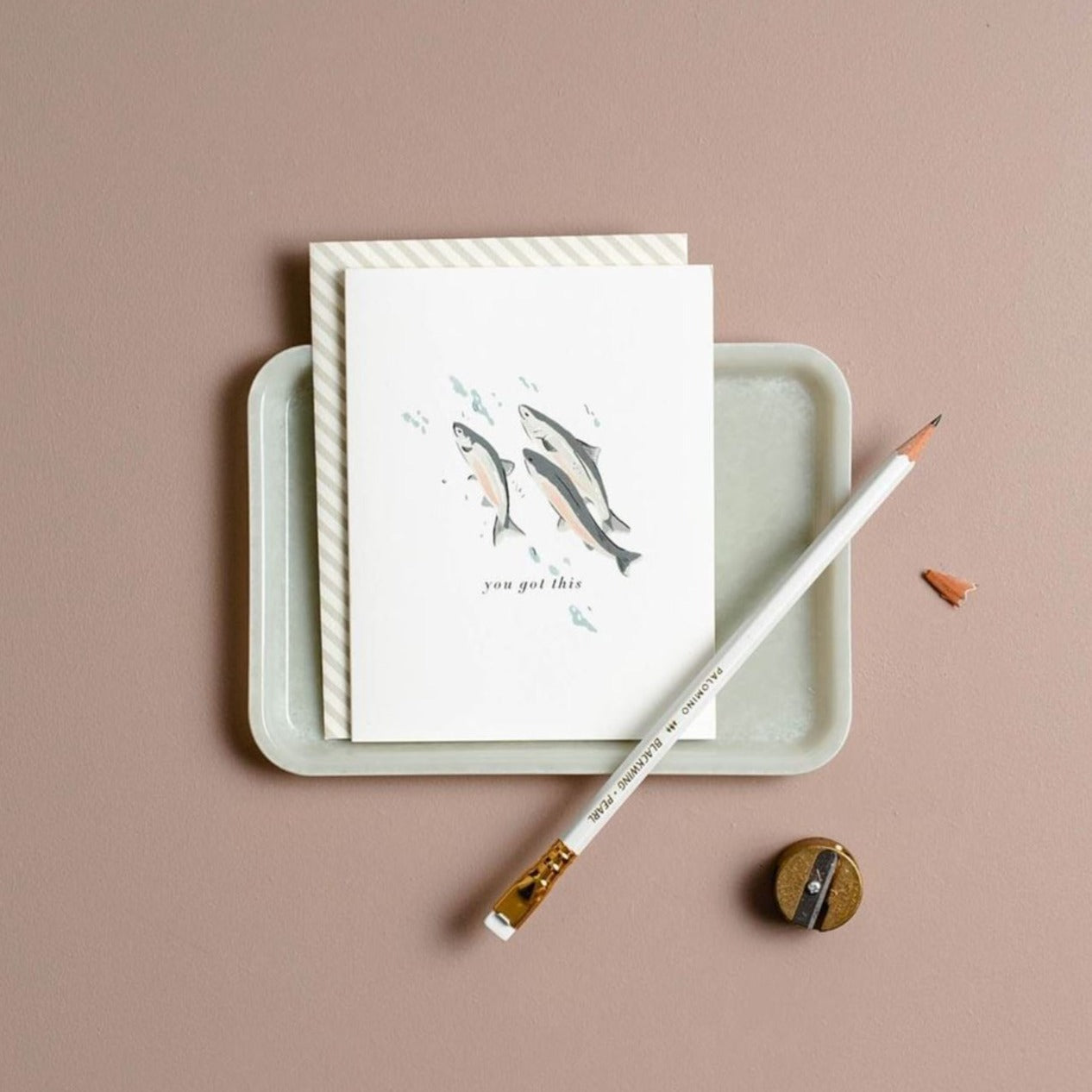 Illustrated 'You Got This' greeting card featuring three fish jumping upstream, displayed on a tray with a pencil and pencil sharpener, all set against a beige background.