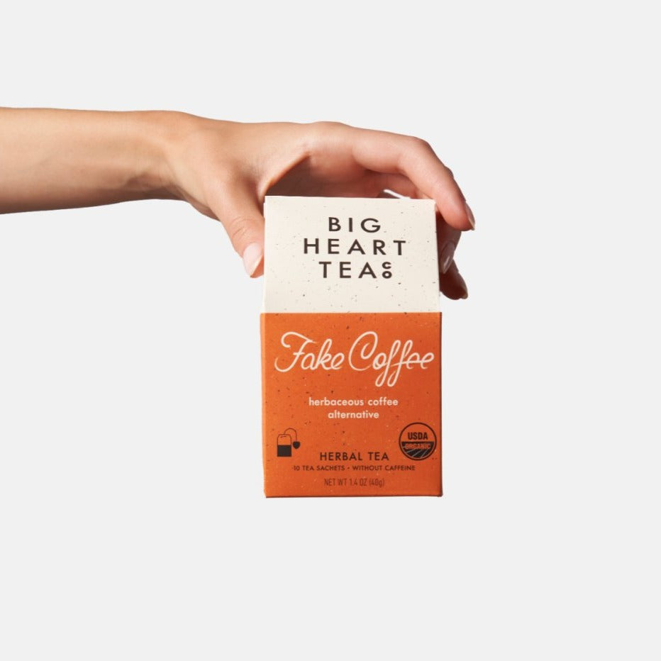 Hand holding a orange box of Fake Coffee, an organic, energizing blend of cacao, roasted chicory, dandelion root, and sweet cinnamon. Touted as 'life-changing' by BuzzFeed, this tea mimics the feel of coffee while offering a chocolaty, herbaceous flavor and natural energy boost.