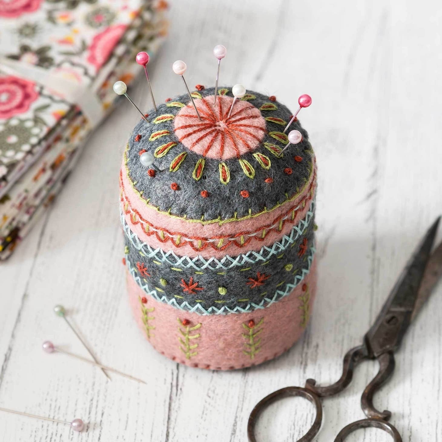 Handcrafted folk-style felt pincushion adorned with intricate embroidery patterns, set against a rustic white wood background, accompanied by vintage scissors and floral fabric.