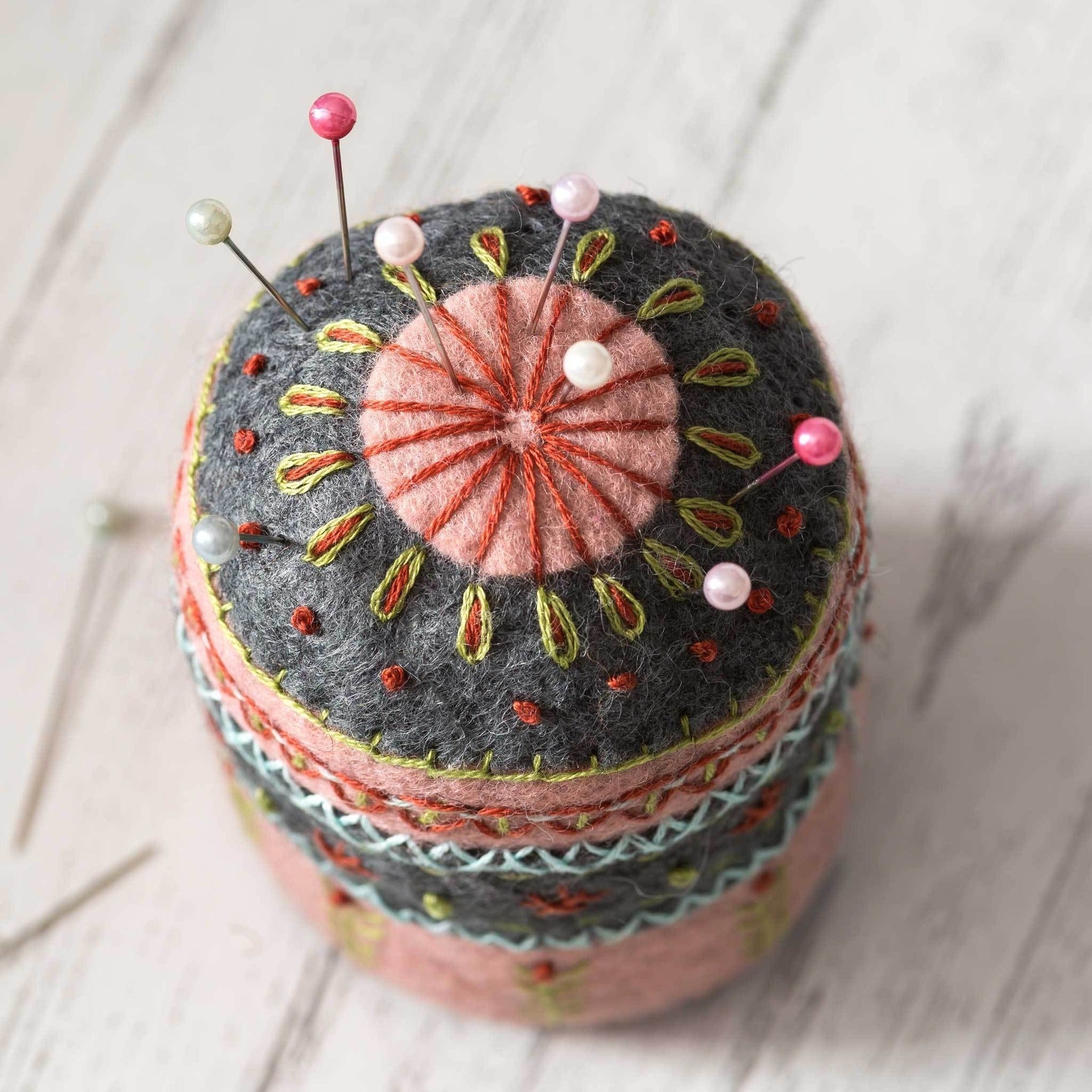 Artisanal felt pincushion with vibrant embroidery details, showcasing a coral sunburst top and colorful stitch patterns, placed on a whitewashed wooden surface.
