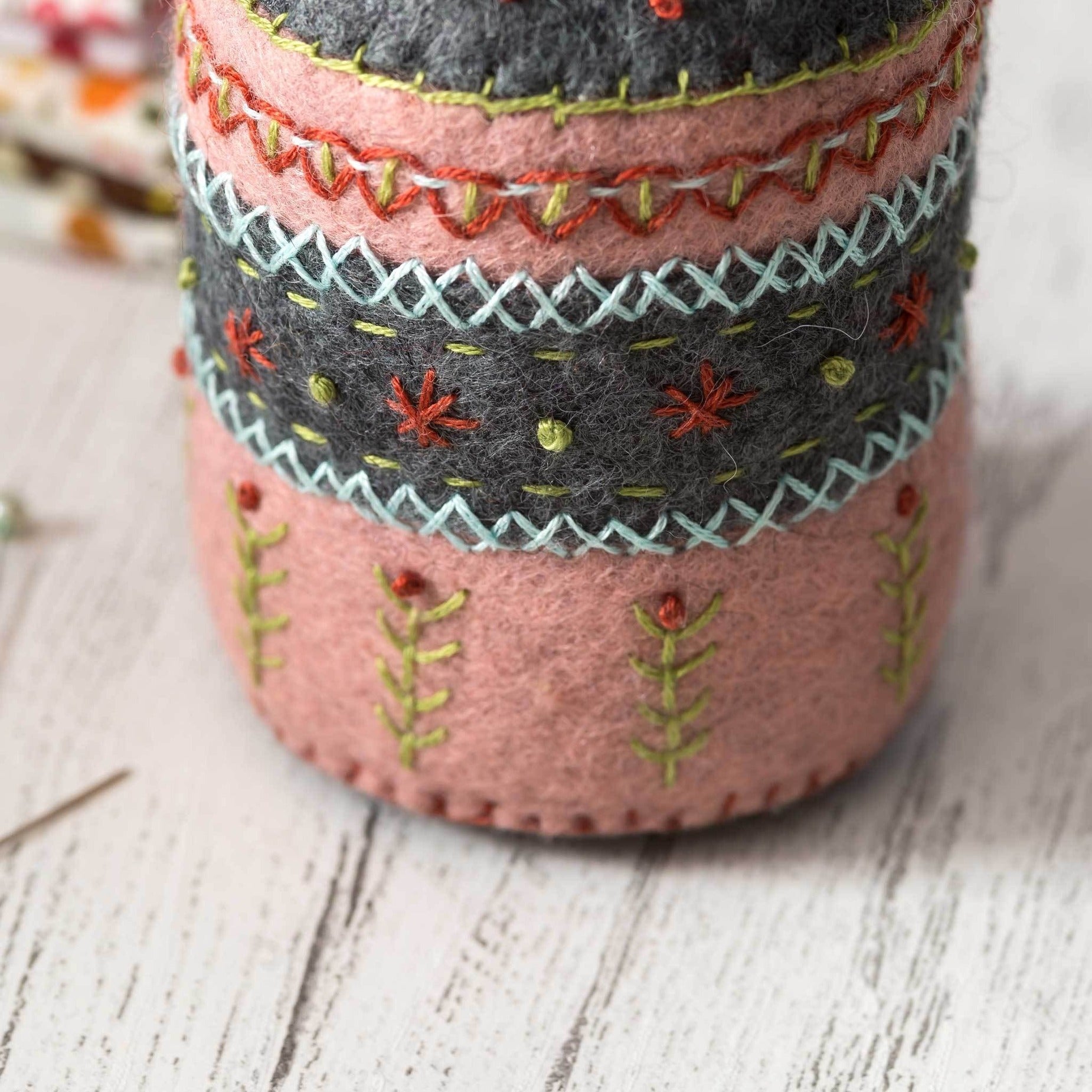 Close-up of hand-stitched felt pincushion layers featuring intricate embroidery patterns in teal, coral, and mustard against a rustic wooden backdrop.