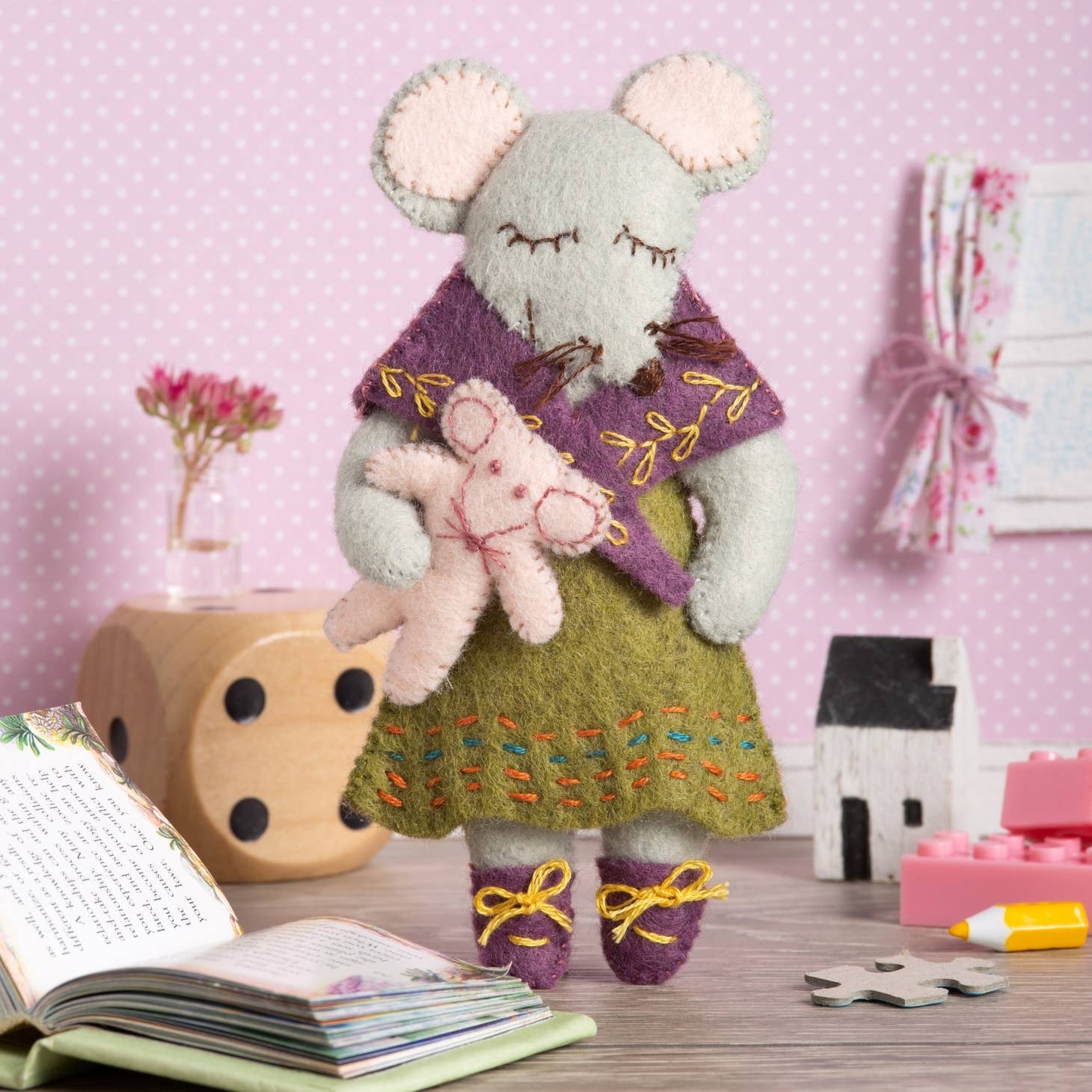 Handcrafted felt mouse figure from the 'Little Miss Mouse Felt Craft Mini Kit', showcasing a delicately dressed mouse holding a soft pink toy against a playful purple polka dot backdrop, surrounded by crafting essentials like a storybook, ribbon-tied fabric, and wooden toys.