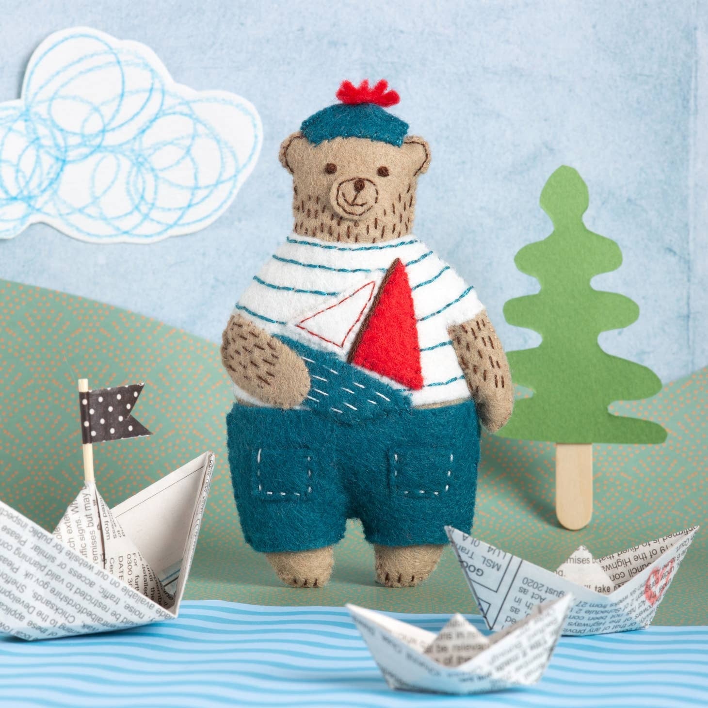 Felted Marcel the Sailor Bear in striped shirt with paper boats and felt trees on a blue background.