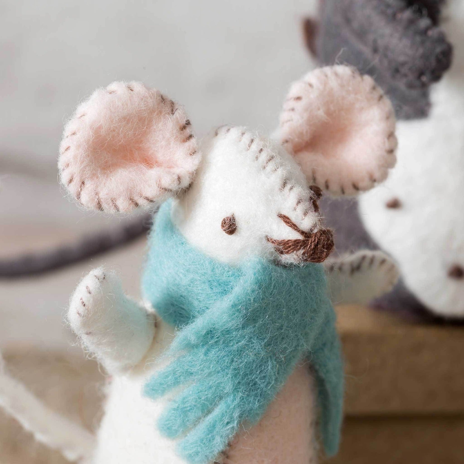 Close-up of a handcrafted white felt mouse with intricate stitching details, wearing a soft teal scarf, set against a muted background with hints of other felt creations.