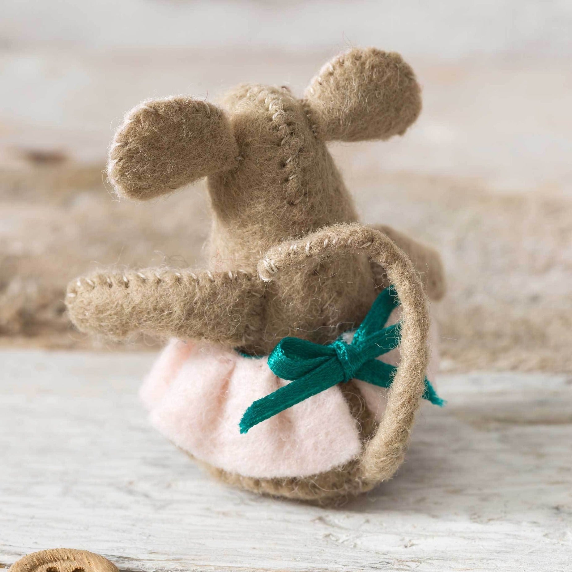 Handmade beige felt mouse, back view, showcasing delicate stitchwork and a pink dress accented with a vibrant emerald green bow, set on a rustic wooden backdrop.