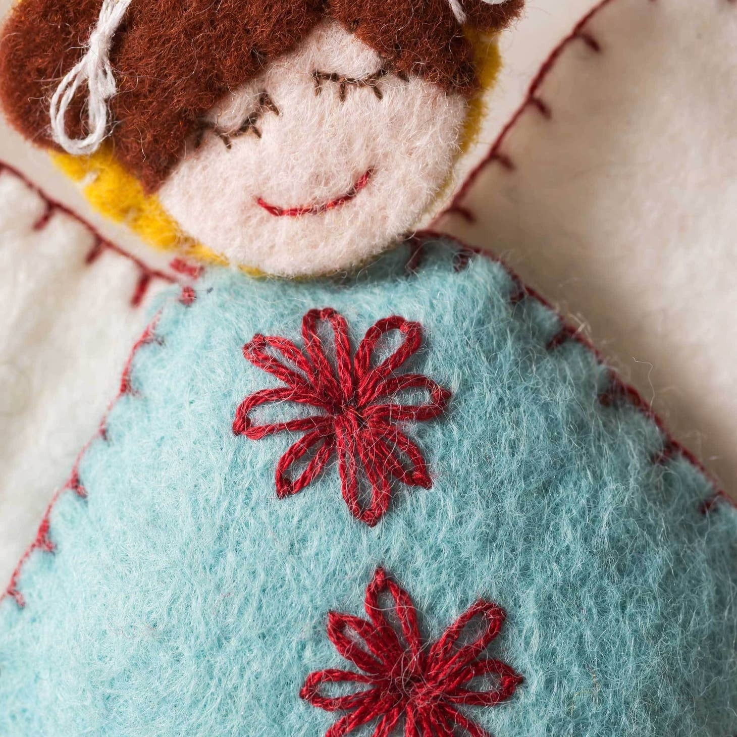 Close-up of a Scandinavian felt angel's smiling face and intricate red embroidered snowflake motifs on soft aqua-blue fabric, showcasing fine craftsmanship and attention to detail.