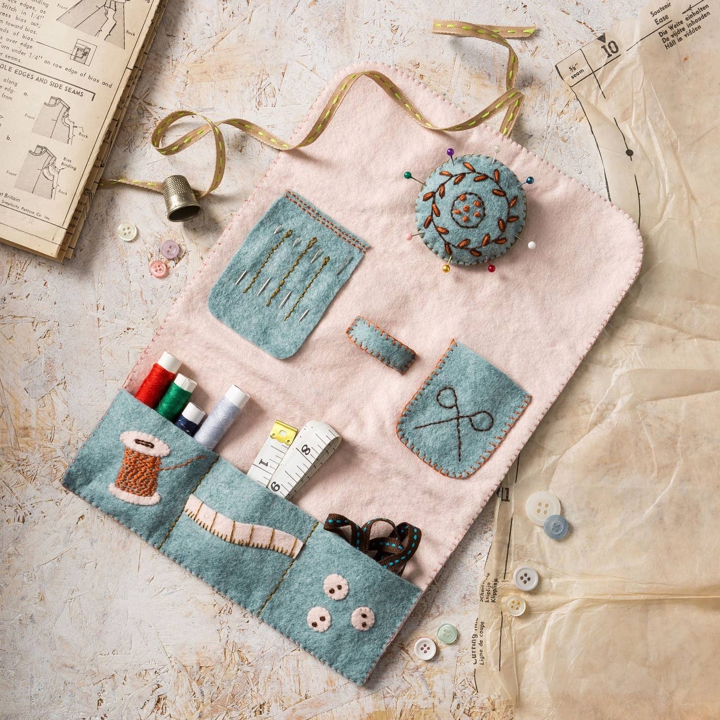 Handcrafted pastel sewing roll kit laid out on a vintage background, showcasing pockets filled with colorful threads, buttons, a tape measure, and a detachable embroidered pin cushion, all bound together with a charming gingham ribbon.