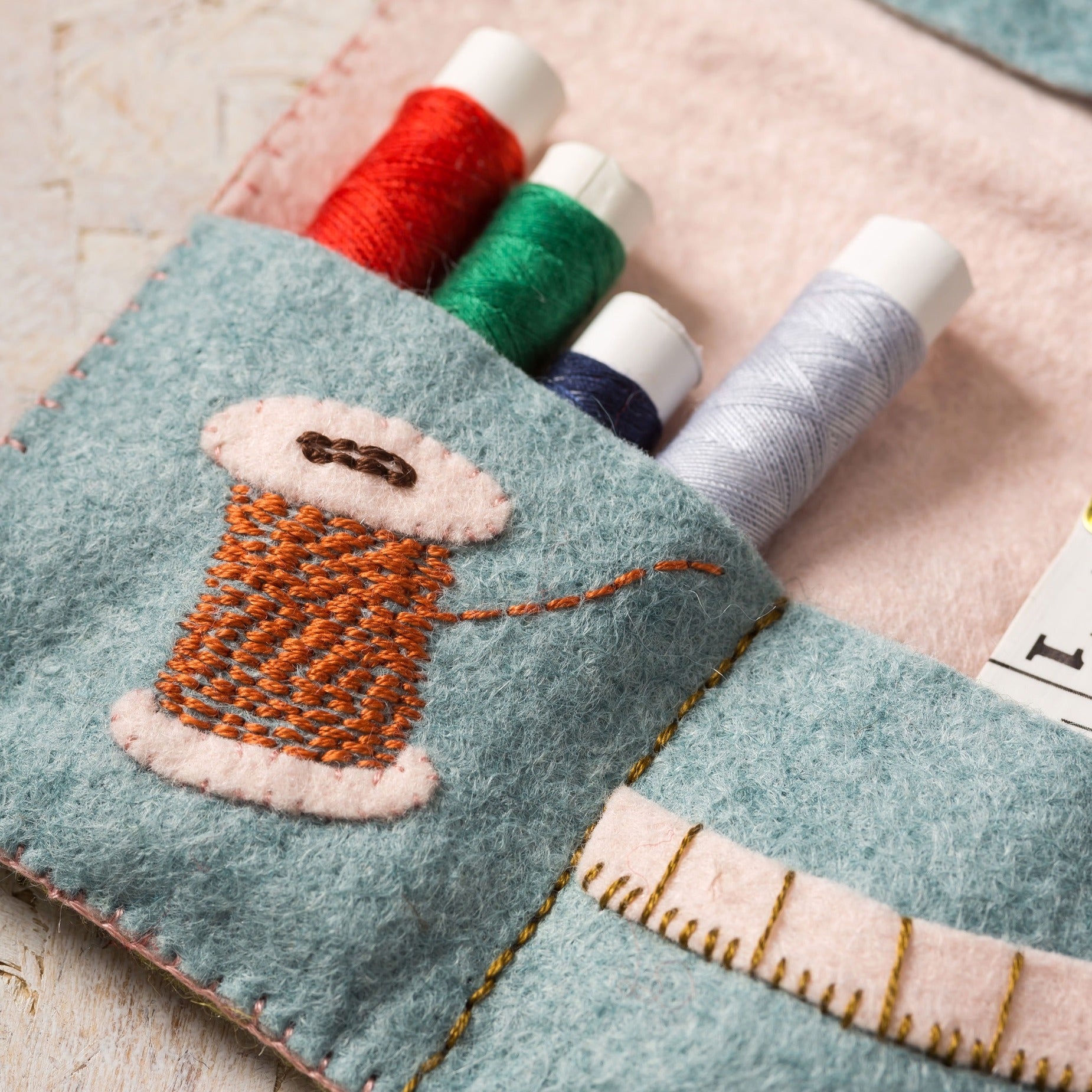 Detail view of a soft teal felt sewing organizer showcasing an embroidered spool of rust-orange thread, accompanied by colorful spools of red, green, blue, and lilac threads on a pink felt background.