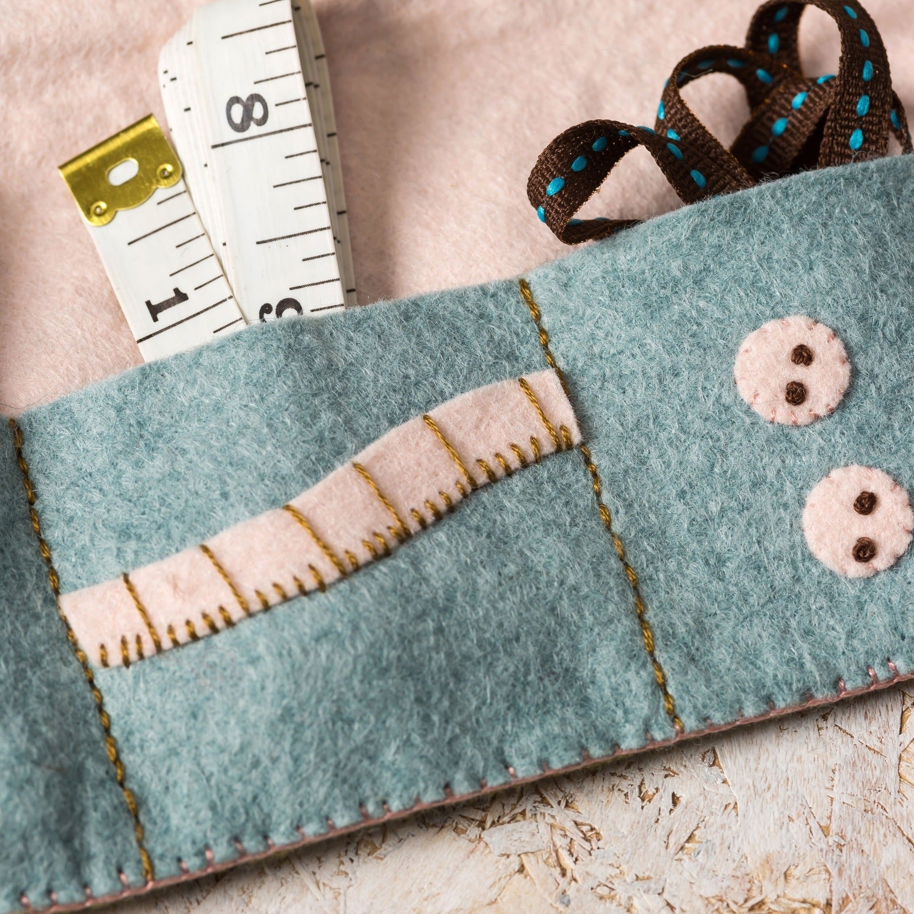 Close-up of a teal felt sewing kit pocket with intricate gold stitching, featuring a vintage white measuring tape and a decorative brown ribbon with blue polka dots, all set on a soft pink felt backdrop.