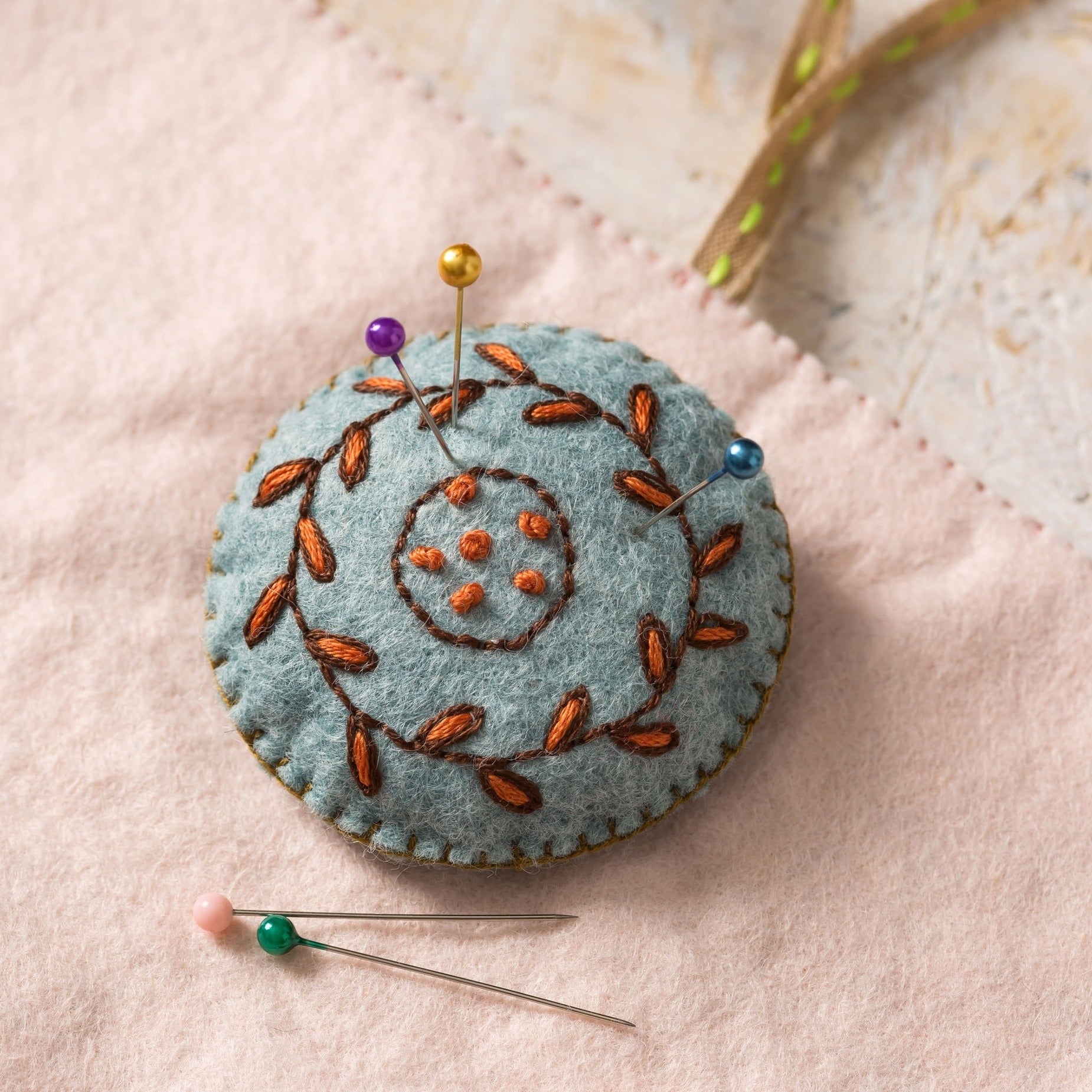 Handcrafted teal felt pin cushion adorned with intricate burnt-orange leaf embroidery and a whimsical button pattern, punctuated with vibrant pins, resting on a soft blush pink fabric background.