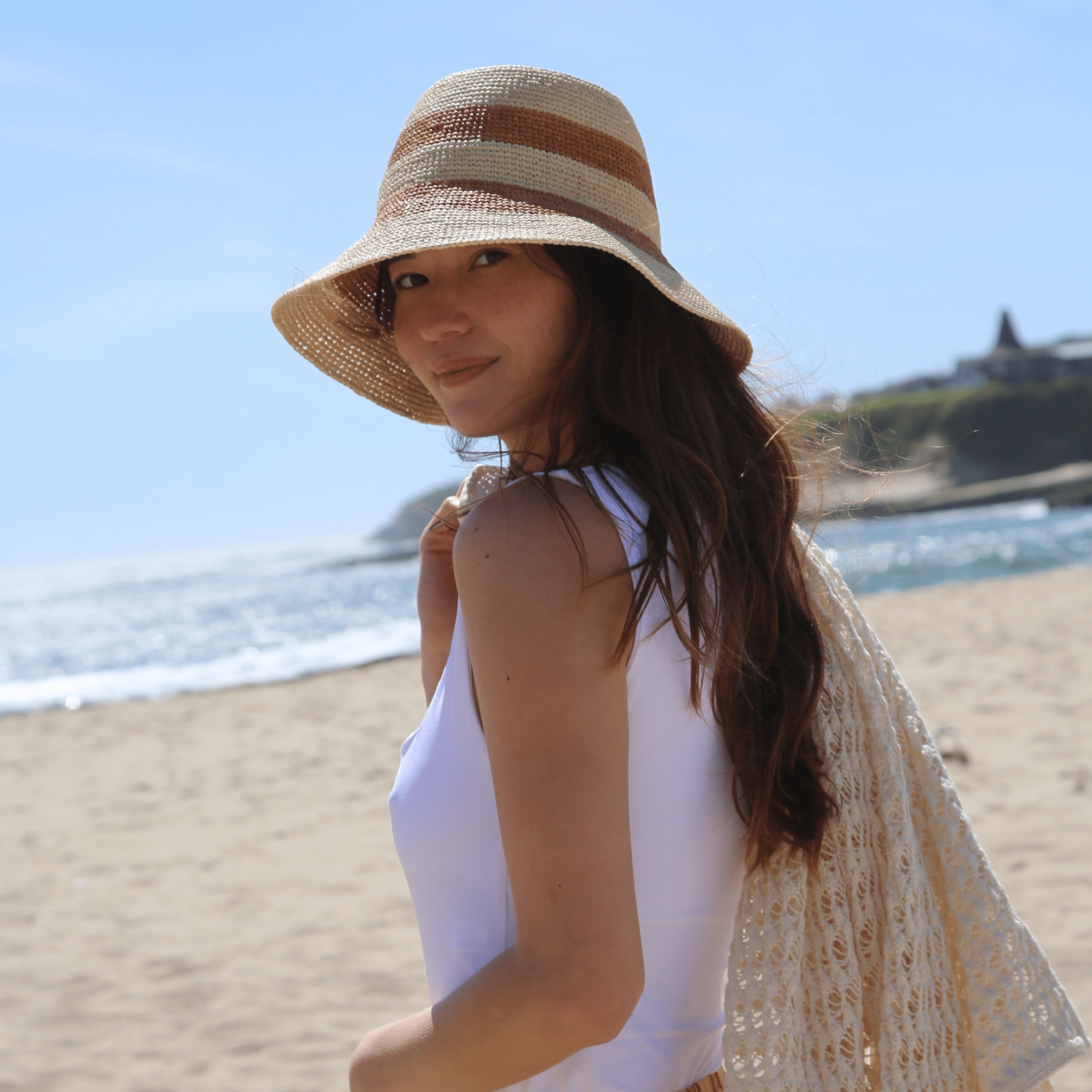 Woman on a beach wearing a straw hat and white top with a beige shawl.
