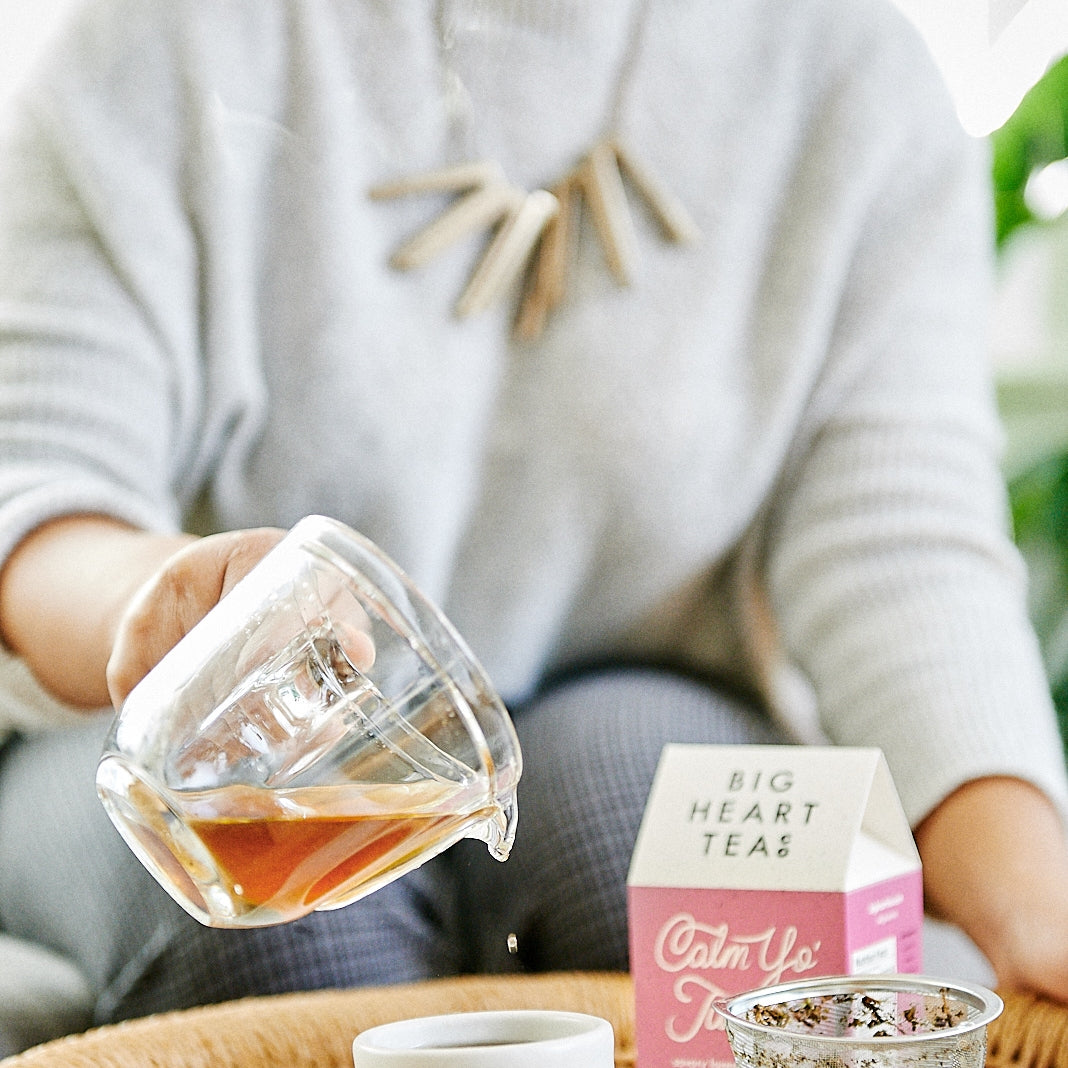 A woman in a gray sweater pouring from a clear mug a cup of Calm Yo' Tummy Tea, an organic blend featuring tulsi, lavender, fennel seed, and brahmi. Designed to stimulate digestion, soothe upset stomachs, relieve stress, and support lactation. Offers a balanced sweet and savory flavor profile.