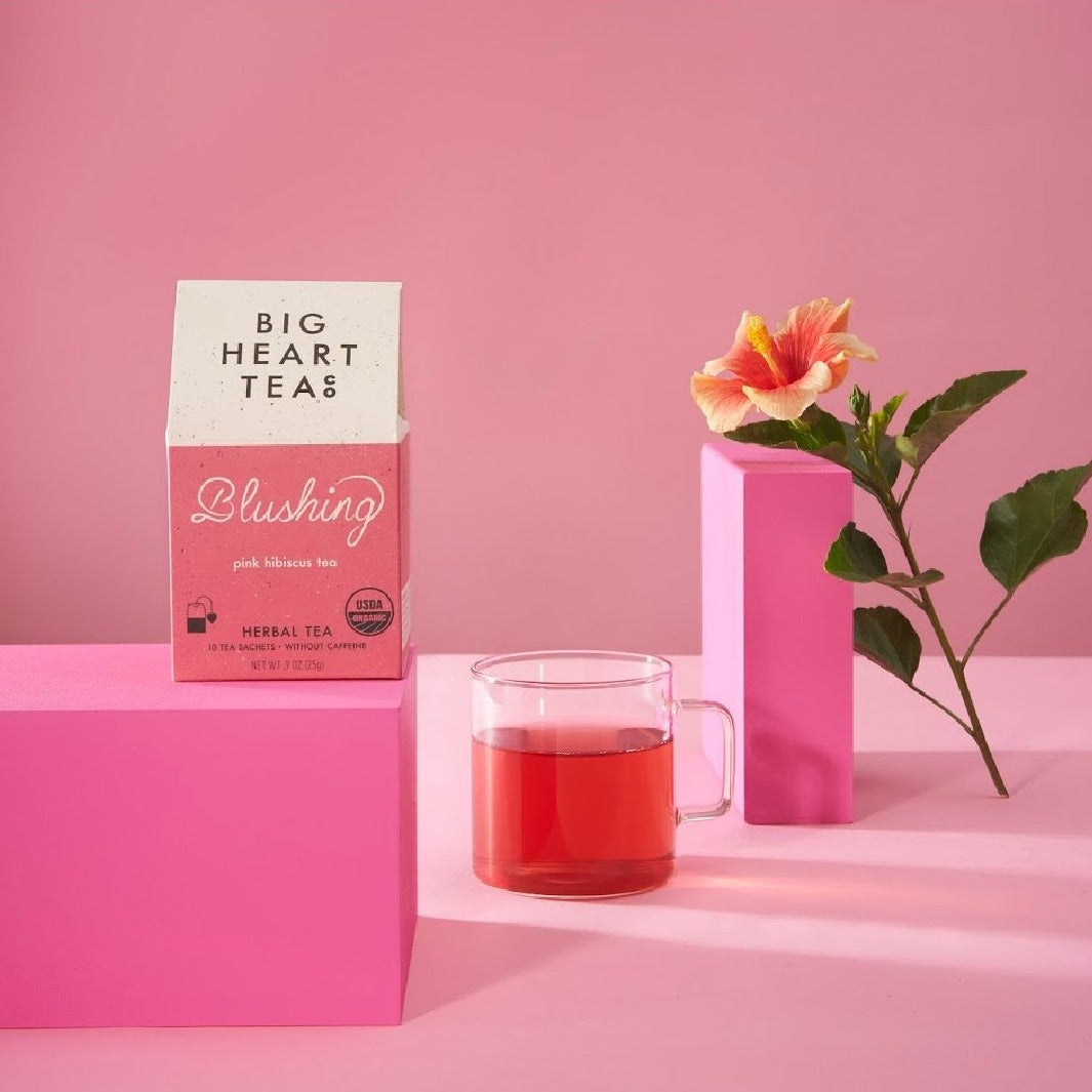 "A pink box of Blushing Tea set on top of a bright pink riser with a hibiscus flower in the background, a naturally caffeine-free blend featuring hibiscus, lemongrass, and tulsi. Ideal for a vitamin C boost and creating a vibrant pink brew that's tart, sweet, and citrusy. Perfect for kids and adults alike.