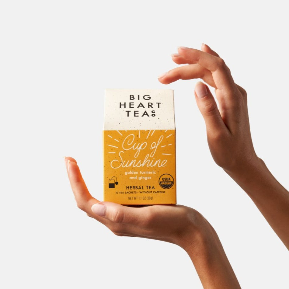 Hand holding Big Heart Tea Co.'s Cup of Sunshine, an organic blend featuring turmeric, ginger, tulsi, Malabar peppercorn, and cinnamon. Known for its anti-inflammatory benefits, immune-boosting properties, and digestive aid.