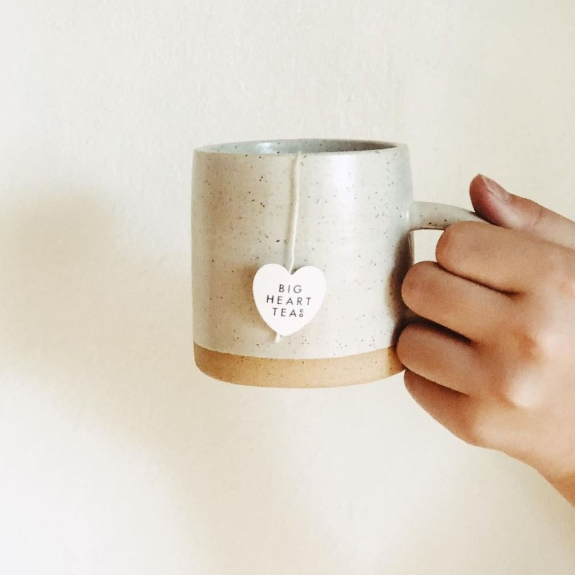 "Hand holding a white ceramic mug with a tea bag of Blushing Tea, a naturally caffeine-free blend featuring hibiscus, lemongrass, and tulsi. Ideal for a vitamin C boost and creating a vibrant pink brew that's tart, sweet, and citrusy. Perfect for kids and adults alike.