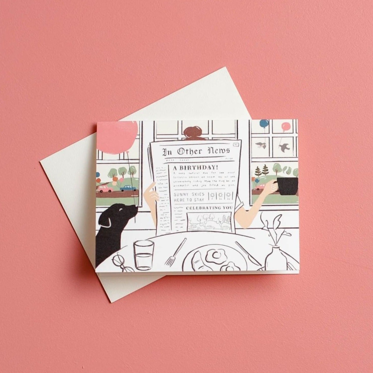 Illustrated chic birthday card on warm white premium cardstock, featuring a woman reading a newspaper at a brunch table with a black lab holding a pink balloon, and a scenic backdrop of birds with balloons through a window.
