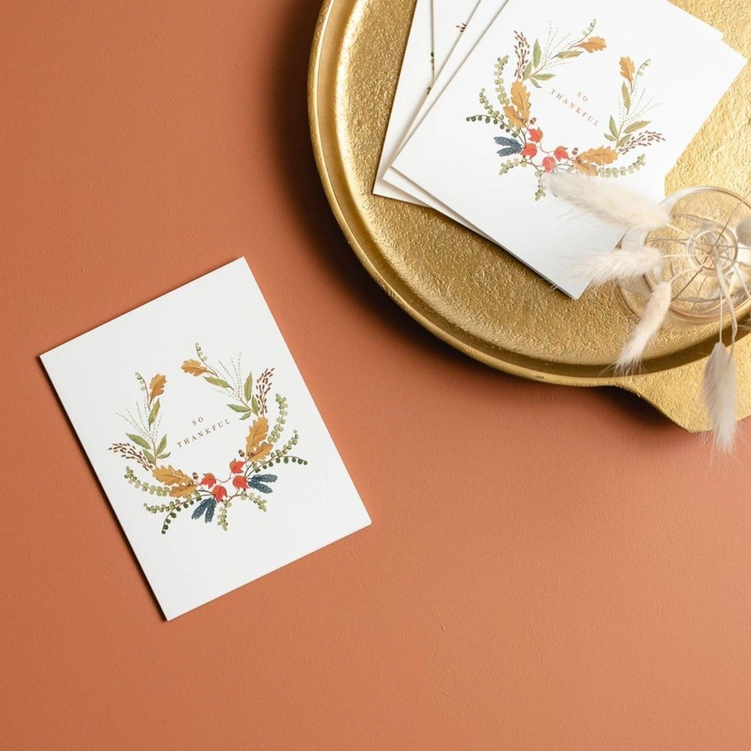 Illustrated Thanksgiving card with a wreath of fall-colored florals surrounding the gold foil words 'So Thankful' on warm white cardstock.