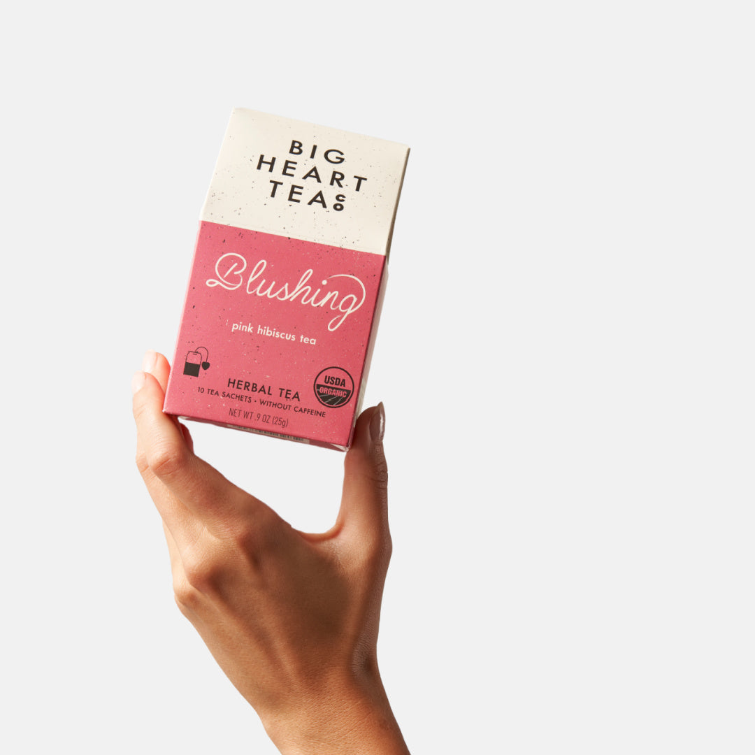 "Hand holding a pink box of Blushing Tea, a naturally caffeine-free blend featuring hibiscus, lemongrass, and tulsi. Ideal for a vitamin C boost and creating a vibrant pink brew that's tart, sweet, and citrusy. Perfect for kids and adults alike.